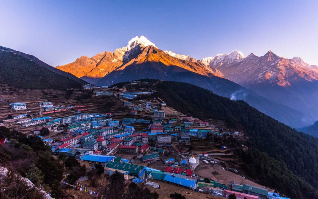 This is the stunning Namche Bazaar, where the mountains are gently kissed by the morning light. The vibrant, multi-colored houses are nestled in the heart of the hills, which rise to embrace both the village and the surrounding peaks, all set beneath a clear blue sky.