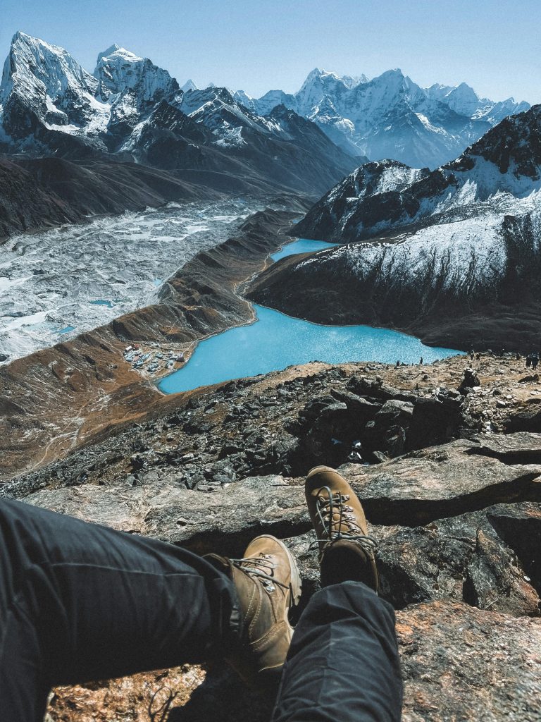 The image shows a breathtaking view of a mountain range with a turquoise lake. The mountains are snow-capped and towering, while the lake is calm and serene.  The perspective is from a hiker's point of view, with their feet resting on a rock. The overall atmosphere is peaceful and majestic