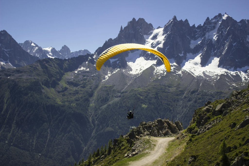 The image captures a thrilling paragliding adventure against a backdrop of majestic mountains. A bright yellow paraglider soars in the clear blue sky, with a person harnessed to it. Below, a winding dirt road climbs a steep mountain slope. The distant mountain range features snow-capped peaks and lush green forests, adding to the scene’s natural beauty. The atmosphere is filled with adventure, freedom, and awe-inspiring views. This picturesque setting could be in a region like the Alps or the Himalayas.