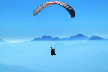 The image shows a paraglider flying high above a beautiful mountain landscape. The paraglider has a bright red and white canopy and is floating in the clear blue sky. Below, a thick layer of clouds stretches out, making a dramatic background. In the distance, tall mountains rise through the mist, their tops hidden in the clouds. The picture captures the peaceful and exciting feeling of paragliding in Pokhara and shows the natural beauty from above.