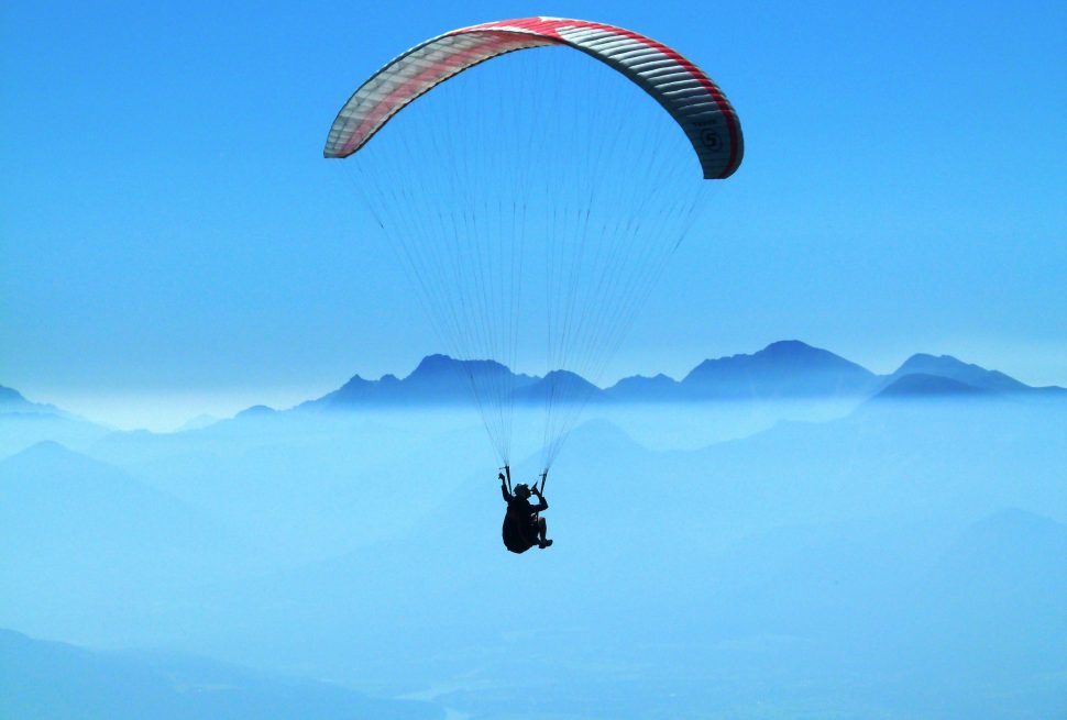 The image shows a paraglider flying high above a beautiful mountain landscape. The paraglider has a bright red and white canopy and is floating in the clear blue sky. Below, a thick layer of clouds stretches out, making a dramatic background. In the distance, tall mountains rise through the mist, their tops hidden in the clouds. The picture captures the peaceful and exciting feeling of paragliding in Pokhara and shows the natural beauty from above.