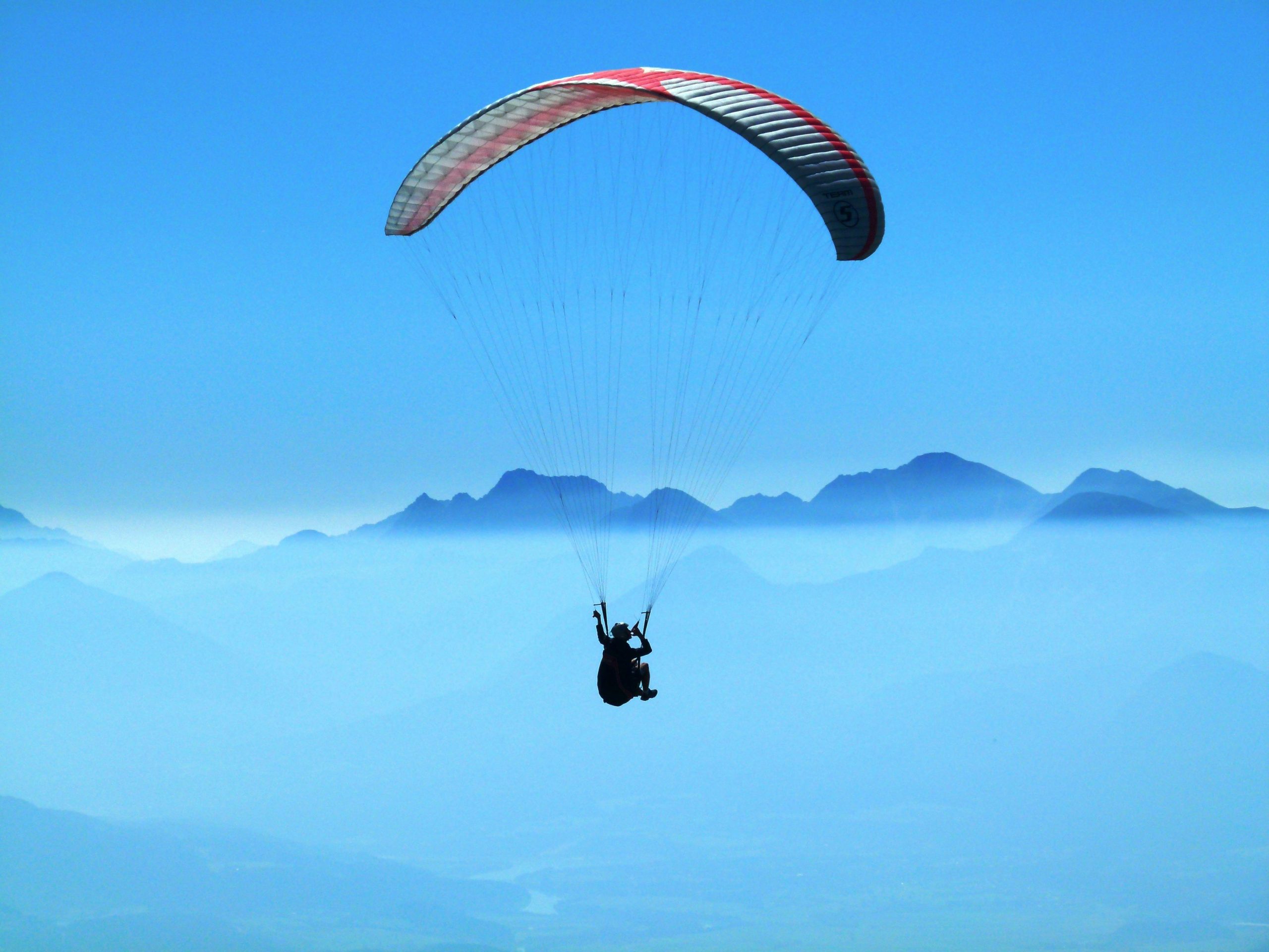 The image shows a paraglider flying high above a beautiful mountain landscape. The paraglider has a bright red and white canopy and is floating in the clear blue sky. Below, a thick layer of clouds stretches out, making a dramatic background. In the distance, tall mountains rise through the mist, their tops hidden in the clouds. The picture captures the peaceful and exciting feeling of paragliding in Pokhara and shows the natural beauty from above.