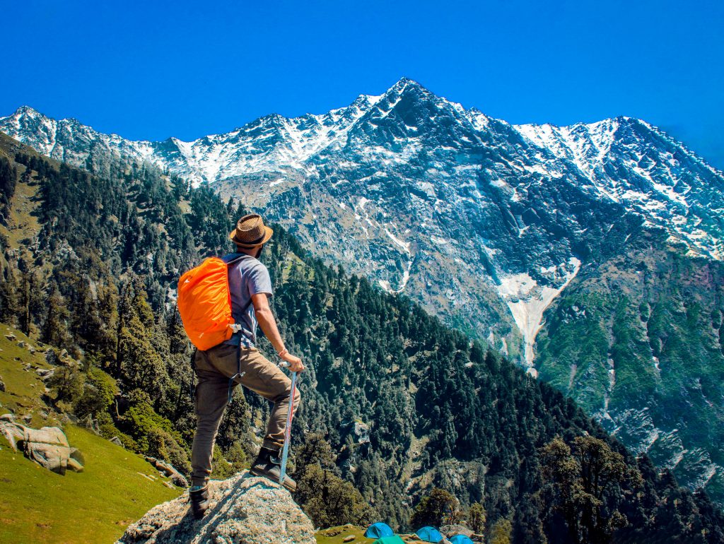 The man is standing on a stone, gazing at the snow-capped mountains and green hills. He is wearing an orange-colored backpack and looking at the landscape, which features various trees, sloping hills, and rocky outcrops.