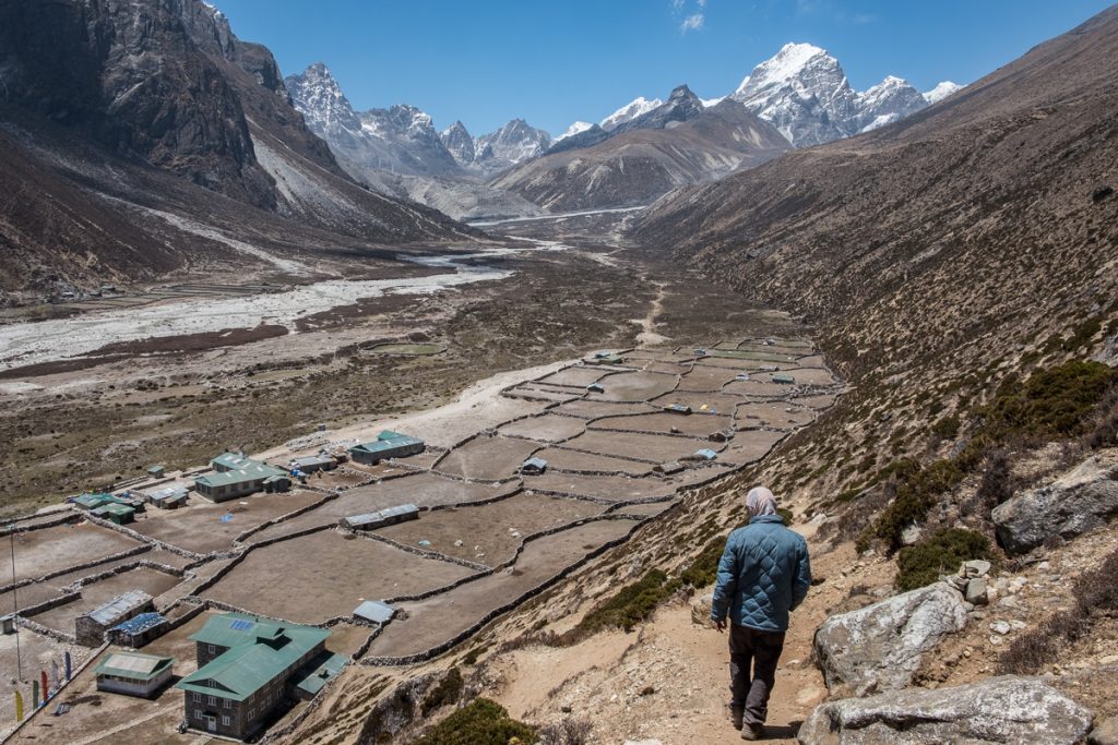 A man stands slightly elevated above the village, gazing down at it. He is dressed in a blue jacket and black pants. Below him, various houses and partially cultivated farmland are visible. In the distance, the majestic Himalayas rise, with green hills extending beside them.