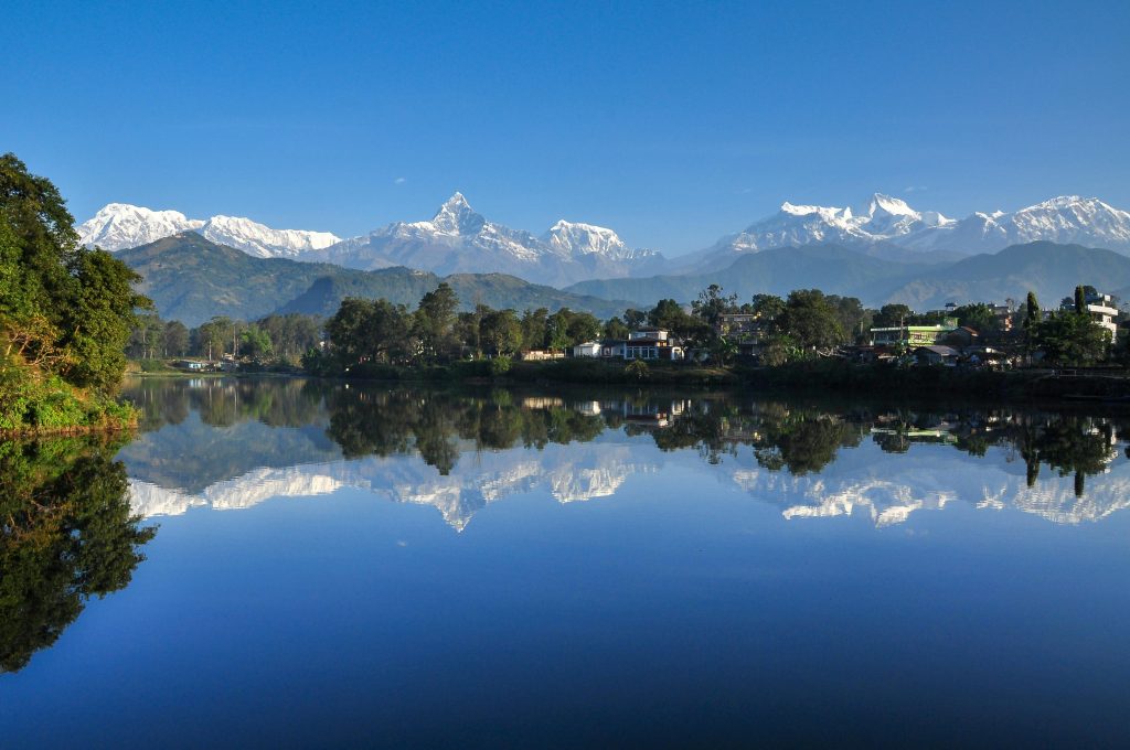 The image captures a breathtaking panoramic view of Phewa Lake in Pokhara, Nepal. The lake's calm, reflective surface mirrors the majestic Annapurna mountain range in the background. The snow-capped peaks of the mountains, including Machhapuchhre and Annapurna South, are reflected in the lake's crystal-clear waters. The foreground features lush greenery, including trees and vegetation, framing the serene scene. The sky is a clear, vibrant blue, completing the perfect picture of natural beauty.