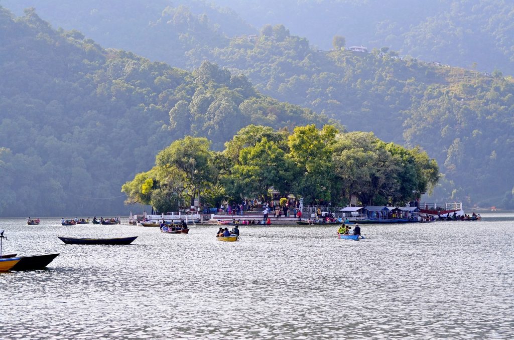 The image shows a calm scene on Phewa Lake in Pokhara, Nepal. In the middle of the lake, there's a small island covered with green trees. On this island, there's a temple, probably Tal Barahi Temple, and colorful boats are carrying people around. The lake is smooth and reflects the distant mountains. The whole scene feels peaceful and highlights the natural beauty of Pokhara.