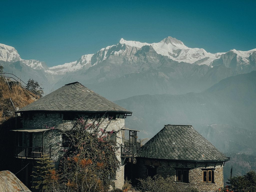 The image shows a beautiful mountainside retreat. Two lovely stone buildings with round roofs are surrounded by green trees. The buildings have traditional Nepalese design with detailed stonework and slate roofs. In the background, there's a stunning snow-capped mountain range. The bright blue sky makes the scene even more serene and beautiful. The picture captures the peaceful and quiet feel of a mountain getaway.