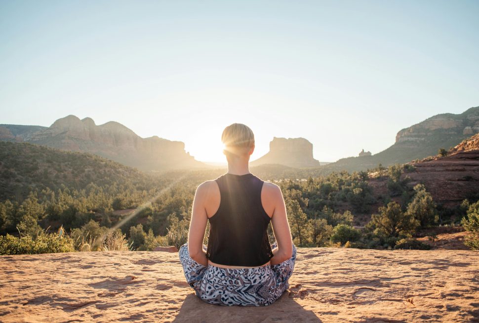 The picture captures a person practicing sunrise yoga on Poon Hill. They are seated on sandy-colored ground, gazing toward the dawn sun, which is peeking through the hills. Between the person and the sun, lush trees create a forest-like frame, adding to the serene, natural beauty of the scene.