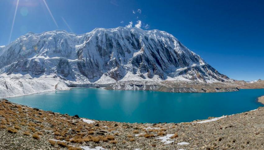 The image depicts a breathtaking landscape featuring Tilicho Lake, one of the highest lakes in the world, nestled amidst the majestic Annapurna mountain range in Nepal. The turquoise waters of the lake, fed by glacial melt, contrast beautifully against the snow-capped peaks that surround it. The sunlight casts a warm glow over the scene, highlighting the rugged beauty of the surrounding terrain. The image captures the serene and awe-inspiring nature of this remote Himalayan region.