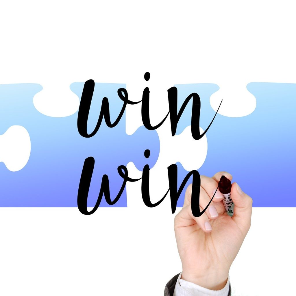 The image shows a hand using a marker to write the word "WIN" twice on a whiteboard. The word is written in the shape of a puzzle piece, with a blue background. This suggests a "win-win" situation, where both sides benefit. The puzzle piece shape symbolizes working together and finding common ground for a positive outcome.