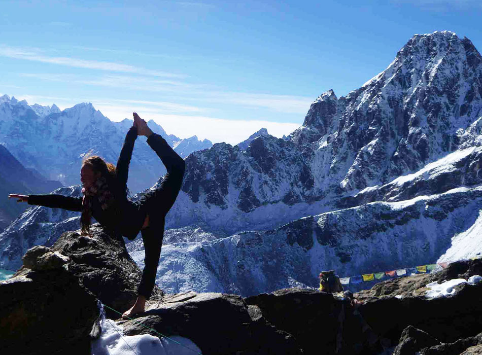 A girl is practicing yoga on a stone, with the snow-capped Himalayas towering behind her. She is surrounded by the majestic mountain range and the clear blue sky.