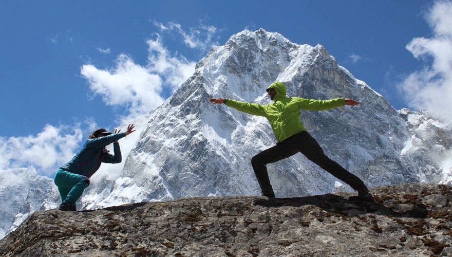 Two people practice yoga in front of the majestic white Himalayas beneath a clear blue sky.