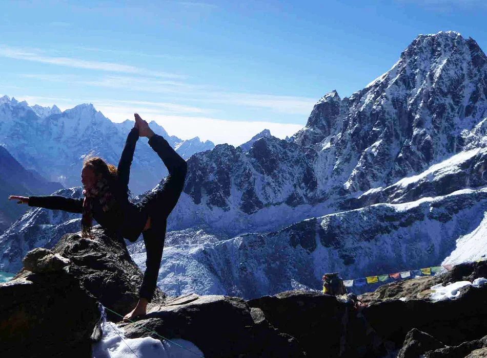 A girl is practicing yoga on a stone, with the snow-capped Himalayas towering behind her. She is surrounded by the majestic mountain range and the clear blue sky.